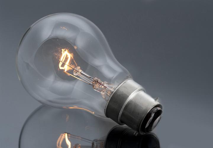 Domestic bayonet fitting light bulb with glowing filament lying on a grey reflective surface in a power and energy concept