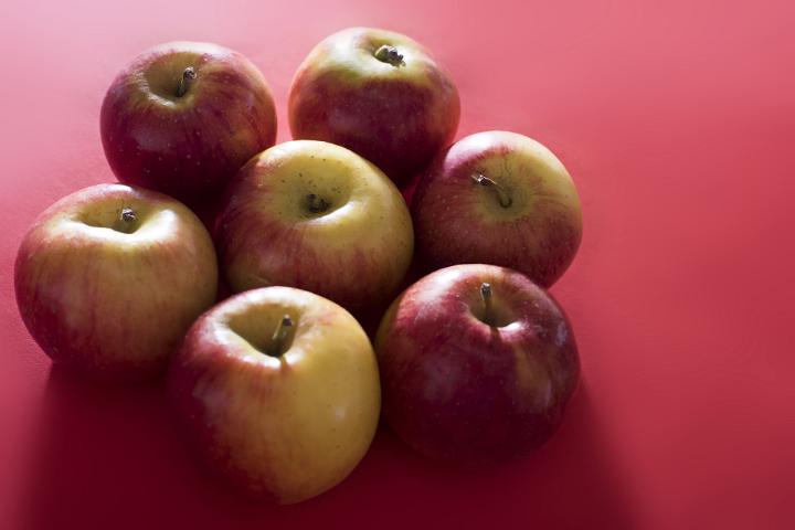 A group of seven apples huddled together lit by bright, contrasting light on a plain red background with copy space.