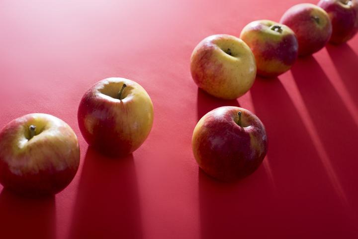 A single odd apple lit by bright light and casting long shadows, sits out of line on a plain red background.