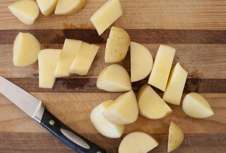 Overhead view of a wooden chopping board and knife being used for dicing and peeling fresh potatoes for cooking as an accompaniment to dinner