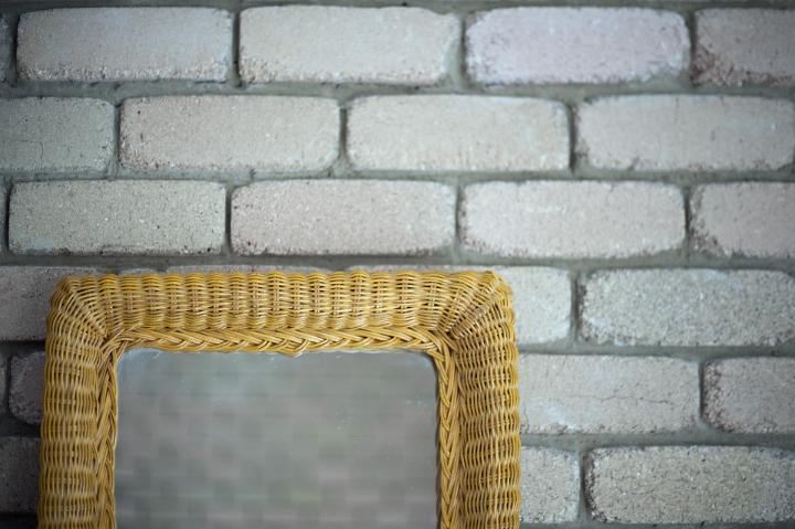 Rattan or wicker framed mirror standing against a rustic white painted brick wall reflecting the brickwork