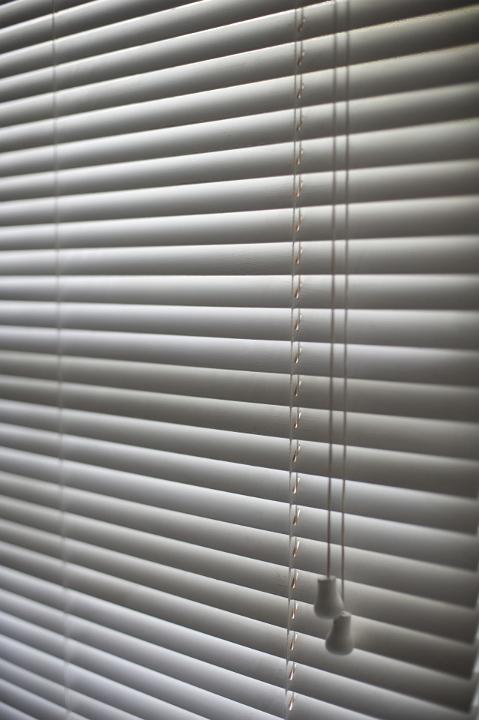 Oblique angle view of grey Venetian blinds hanging on a window with the slats closed and toggles for opening and closing in the foreground