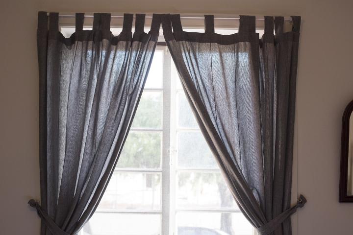Window in a home with dark colored drapes or curtains hanging on a simple rod pulled to the sides with ties in a translucent fabric