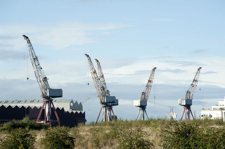 Row of large industrial cranes in a ship building yard standing on the wharf in front of a large warehouse at a port