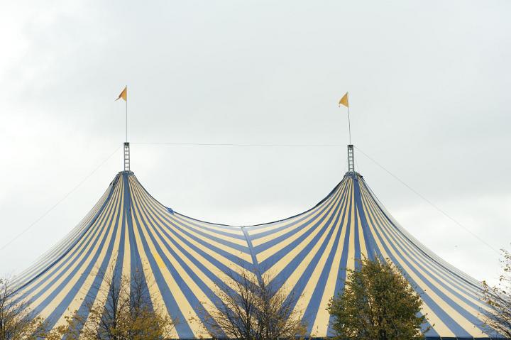 Colorful top of a striped marquis for a music festival visible above a row of green trees against a cloudy blue sky