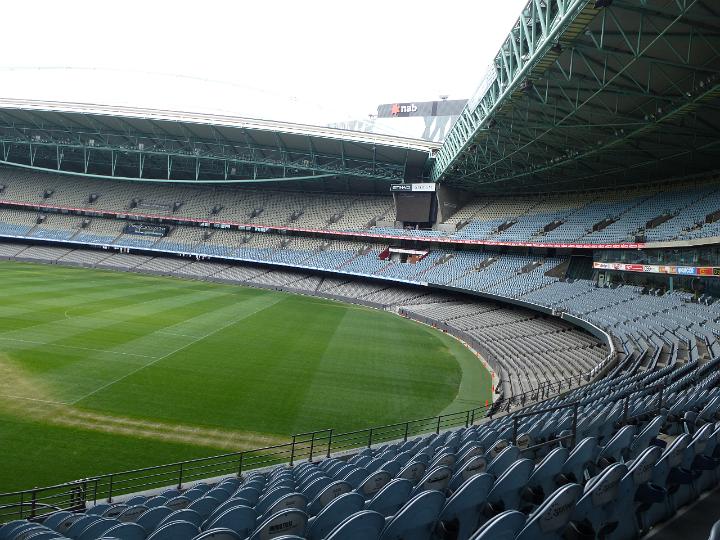 Empty tiers of seating in a sports stadium oval around a green grassy sports field with a view of curved seating at the top of the field