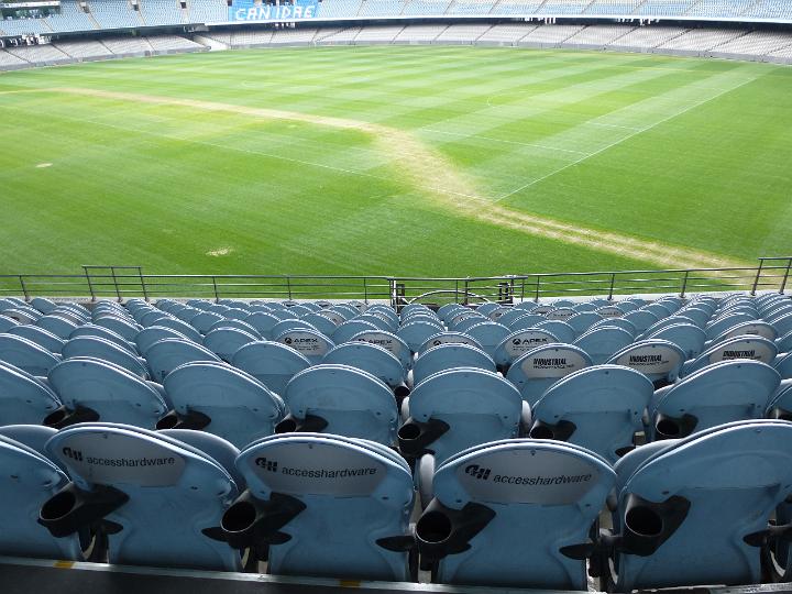 Empty seating at a sports stadium viewed from behind looking down towards the green field below