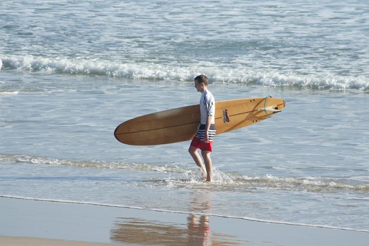 Surfer walking along the waters edge on the beach sand carrying his surfboard under his arm, side view on a sunny summer day